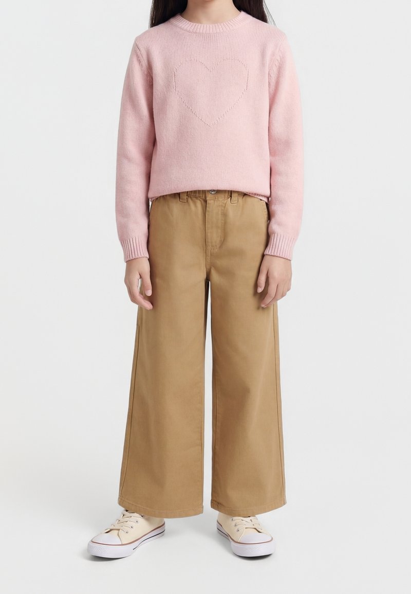 Enfant portant un pull rose avec un motif de cœur, un pantalon large beige et des baskets couleur crème, debout sur un fond blanc.