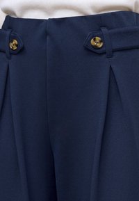 Gros plan sur un pantalon bleu marine avec des plis et des passants décoratifs maintenus par des boutons beige à la taille.
