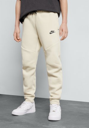 Pantalones de chándal beige de Nike con una cintura ajustable, que presentan un logo negro en el muslo izquierdo y detalles de paneles laterales texturizados. Llevados con zapatillas blancas.