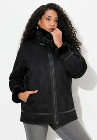 Giacca in finta suede nera con colletto alto, chiusura con zip, tasche laterali e polsini morbidi. Presenta sottili strisce orizzontali lungo l'orlo.