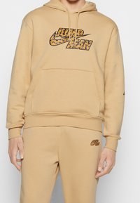 Sudadera con capucha color canela con un gran logo estampado de animal y detalles en negro. Cuenta con un bolsillo frontal y puños acanalados. Textura suave, ajuste informal.