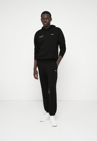 Czarna bluza z kapturem z przednią kieszenią i haftowanym logo, w połączeniu z pasującymi czarnymi dresami. Całość dopełniają białe sneakersy.