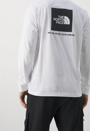 Personne portant une chemise blanche à manches longues avec le logo noir "The North Face" et le texte "Never Stop Exploring" dans le dos, ainsi qu'un pantalon noir.