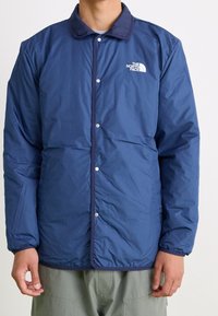 Blå isolerad jacka med tryckknappsstängning, utrustad med krage och North Face-logotypen på vänster bröst. Elastiska ärmavslut och slät textur.