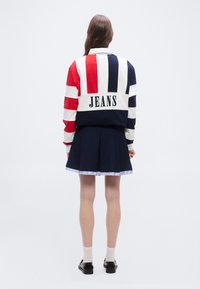 Άτομο με μακριά καστανά μαλλιά, φοράει κόκκινο, λευκό και μπλε ριγέ σακάκι με την επιγραφή "JEANS", ναυτικό πλισέ φούστα, λευκές κάλτσες και μαύρα παπούτσια.