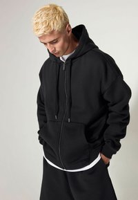 Black zip-up hoodie made from thick fabric. Features a hood, front pockets, and ribbed cuffs. Worn over a white t-shirt and pants.