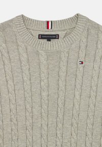 Jasnoszary sweter z dzianiny z wzorem w warkocze, ściśle przylegającym do szyi i małym logo flagi na piersi. Miękka tekstura, klasyczny krój.