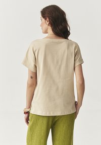 Beige pamut póló rövid ujjakkal, széles nyakkivágással és enyhén ívelt aljjal, texturált zöld nadrággal párosítva.
