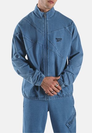 Mann trägt eine blaue Reebok-Jacke mit Reißverschluss und passende Hose, steht mit vor dem Körper verschränkten Händen vor neutralem Hintergrund.