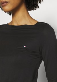 Primo piano del collo e della spalla di una persona che indossa un top nero con un piccolo logo colorato sul lato sinistro, evidenziando il tessuto testurizzato.