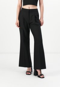 Woman wearing black high-waist wide-leg trousers and black heeled sandals against a plain white background.