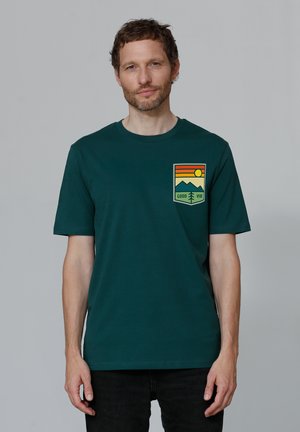 Donkergroen katoenen T-shirt met korte mouwen, voorzien van een grafische zak aan de voorkant in oranje, geel en groen, die bergen en de zon afbeeldt.