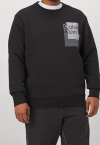 Schwarzer Pullover mit Rundhalsausschnitt, mit einem grauen rechteckigen Logo-Patch, auf dem "Calvin Klein" in fetten schwarzen Buchstaben steht.
