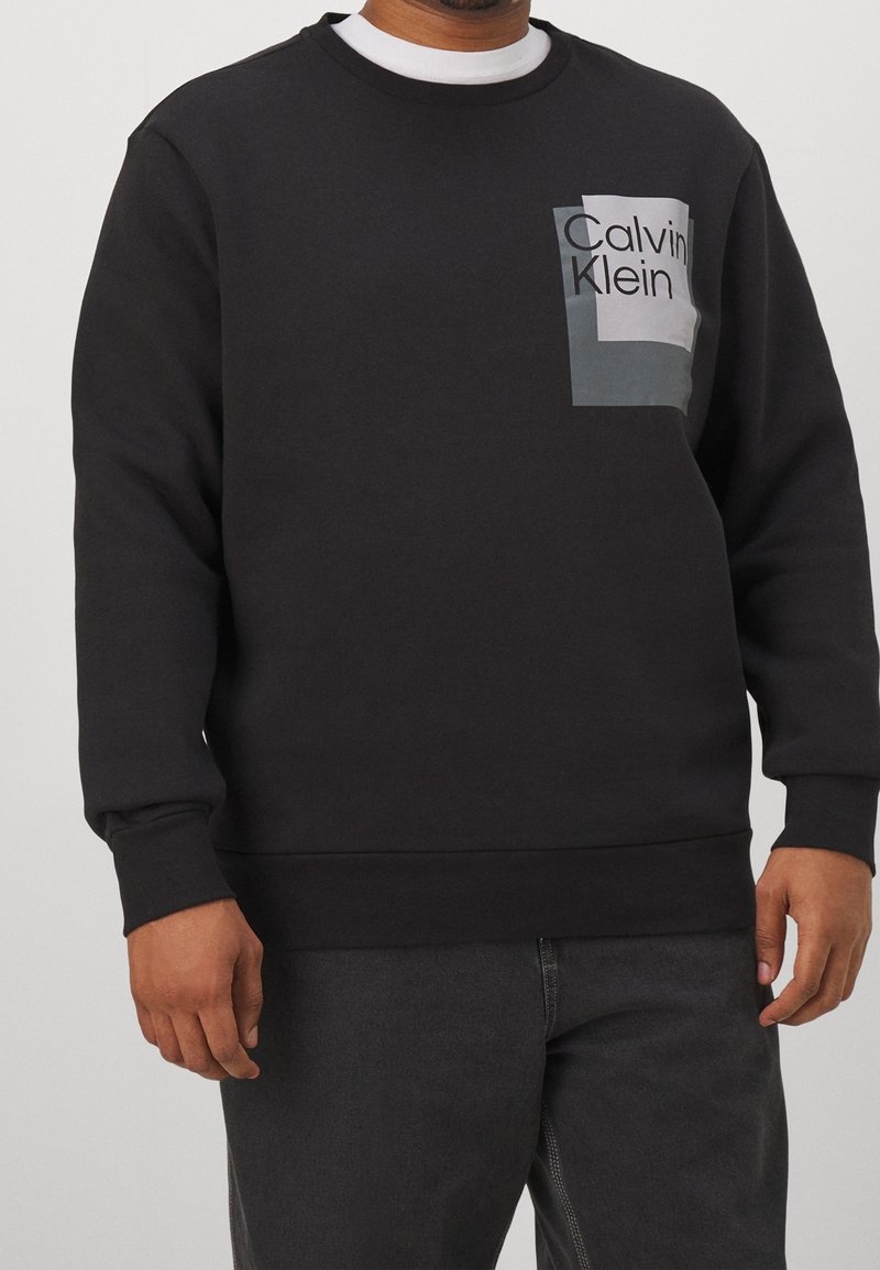Schwarzer Pullover mit Rundhalsausschnitt, mit einem grauen rechteckigen Logo-Patch, auf dem "Calvin Klein" in fetten schwarzen Buchstaben steht.