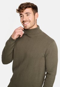 Zeleni pleteni pulover s visokim ovratnikom koji sadrži teksturirani riblji kost pattern i rebraste manžete i ovratnik. Glatka tkanina, usklađen dizajn.