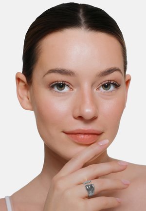 Młoda kobieta z gładką skórą i zielonymi oczami, trzymająca podbródek ręką noszącą srebrny, wyrazisty pierścień, neutralne tło.