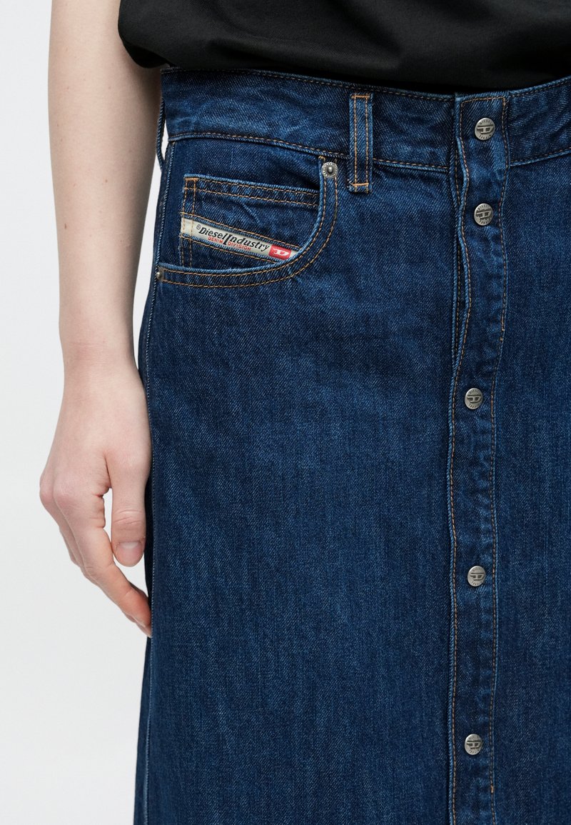 Denim skirt with a dark blue hue, featuring a front button closure and side pockets. Visible brand tag in the back pocket.