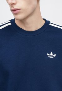 Μπλε ναυτικό φούτερ από βαμβάκι, με στρογγυλή λαιμόκοψη, λευκό σχέδιο με τρεις ρίγες στους ώμους και μικρό λευκό λογότυπο στο στήθος.