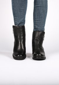 Botas de cuero negras hasta el tobillo con acabado texturizado, cremallera lateral, punta redondeada y tacón bajo, llevadas con jeans de mezclilla azules.