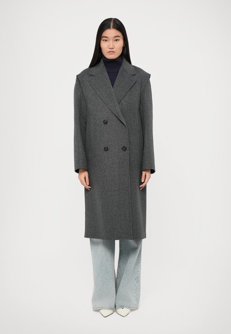 Cappotto di lana grigio a doppio petto con ampi rever e bottoni neri, abbinato a jeans larghi blu chiaro e scarpe bianche a punta.