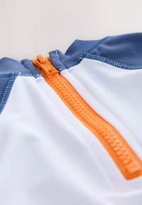 Kurzärmeliges Oberteil mit weißem Körper, blauen Akzenten und einem orangefarbenen Reißverschluss vorne. Glatte Textur mit kontrastierenden Nahtdetails.