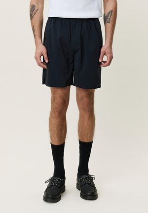 Mann trägt schwarze Shorts, schwarze Socken, schwarze Schuhe, weißes T-Shirt, mit Tattoos auf den Unterarmen, steht vor einem schlichten hellen Hintergrund.