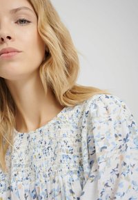 Bluzka w kwiatowy wzór w kolorze białym, z niebieskimi i pastelowymi akcentami, z marszczoną halką i bufkami, wykonana z lekkiego materiału.