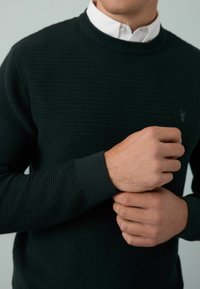 Uomo che regola il polsino di un maglione verde scuro a trama sopra una camicia bianca su uno sfondo semplice.