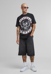 Juodas grafikos marškinėliai su „Motorhead“ dizainu. Sujungti su tamsiai mėlynomis džinsinėmis šortais ir baltomis sportinėmis bateliais. Aksesuarai apima saulės akinius ir tatuiruotes.