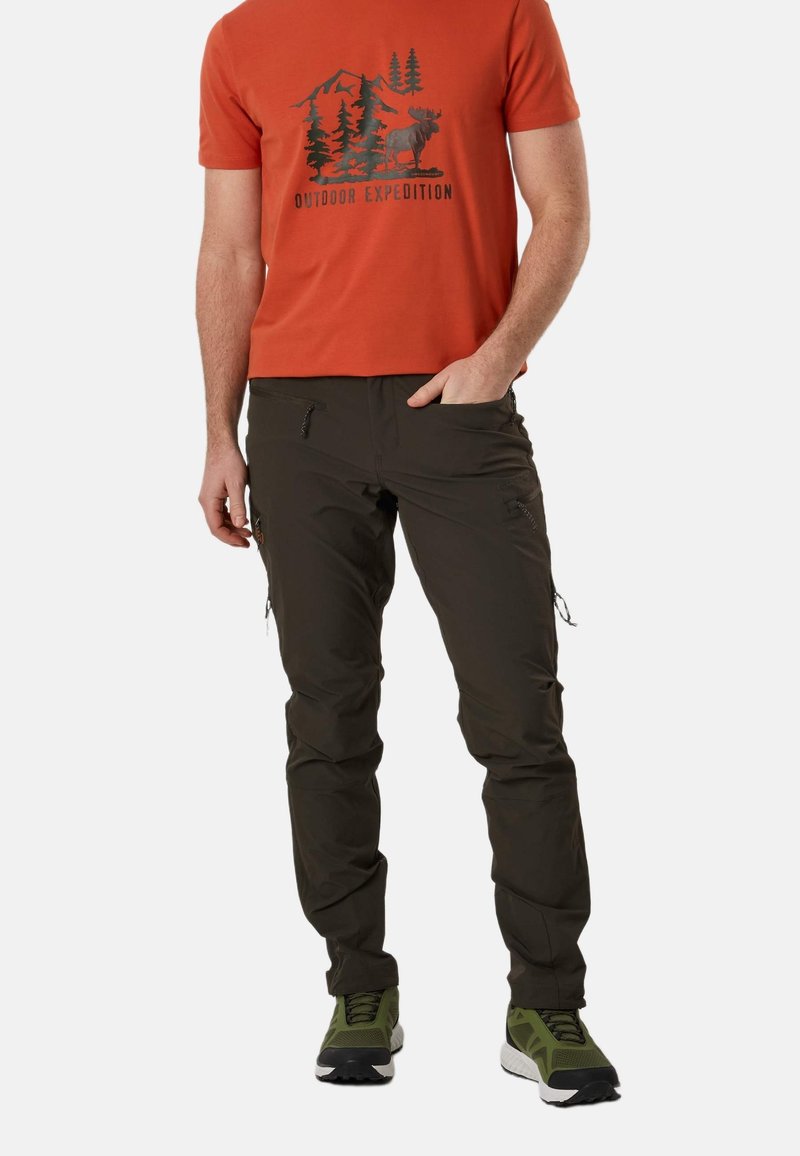 Homme portant un t-shirt orange avec un motif extérieur, un pantalon cargo foncé et des chaussures de randonnée vertes, debout avec une main dans la poche.