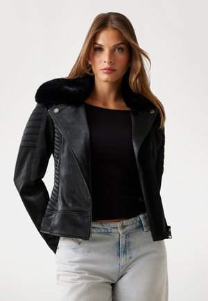 Giubbotto da moto in pelle nera con colletto in pelliccia sintetica, dettagli a costine sulle maniche e design aderente. Abbinato a jeans azzurri.