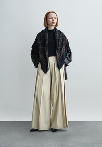 Fekete sál geometriai mintával, fekete pulóverre rétegezve, krém színű, bő szárú nadrággal. A megjelenést fekete cipő egészíti ki.