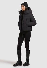 Sort pufferjakke med hætte, der har en lynlås og trykknapper. Båret over en sort top og leggings, parret med knæhøje støvler.