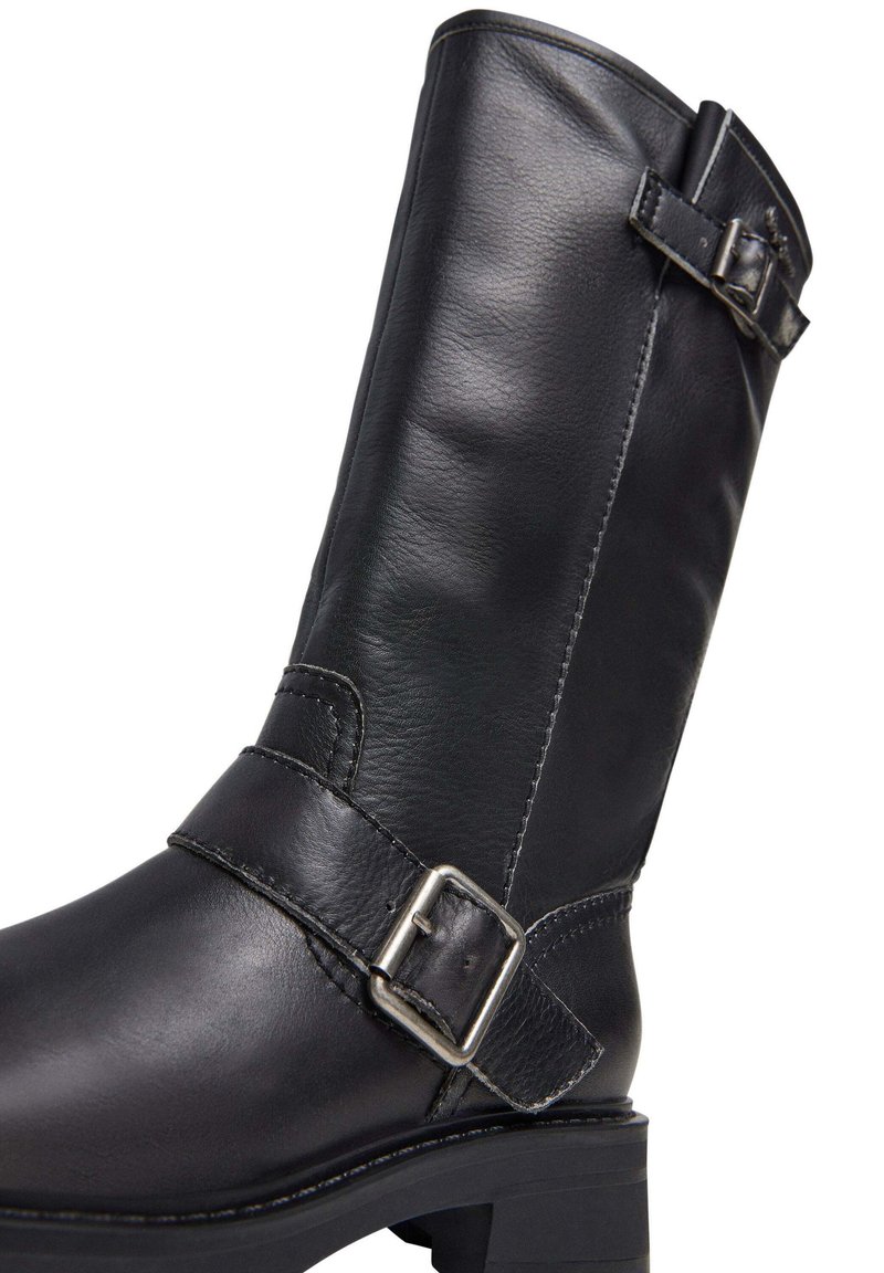 Stivale in pelle nera fino al ginocchio con punta arrotondata, dettagli con fibbie laterali e finitura texturizzata. Presenta un tacco basso a strati e cuciture a contrasto.