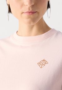 T-shirt di cotone rosa chiaro con scollo rotondo, dotato di un piccolo logo geometrico dorato sul lato sinistro del petto. Texture liscia, design semplice.