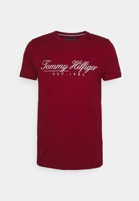 Rød bomulds T-shirt med korte ærmer, med hvid kursiv tekst "Tommy Hilfiger EST. 1985" på tværs af brystet. Ren design, rund halsudskæring.