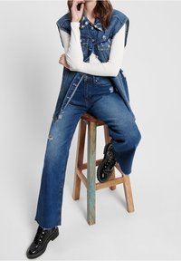 Denimväst med slitna detaljer, lagd över en vit långärmad topp. Vida blå jeans tillsammans med svarta stövlar. Bakgrund av en träpall.