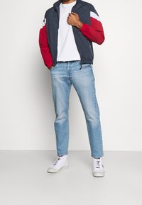 Urheilullinen tummansininen takki, jossa on punaiset ja valkoiset raidat, yhdistettynä vaaleansinisiin farkkuihin ja valkoisiin lenkkikenkiin. Rentoa, mukavaa istuvuutta ja sileä rakenne.