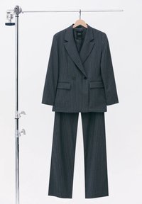 Blazer gris foncé à rayures fines et pantalon à jambes larges assorti suspendus sur un porte-vêtements en métal avec un cintre en bois.