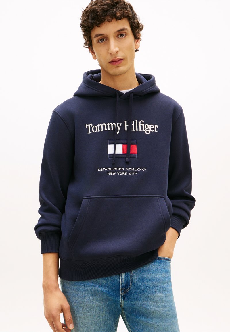 Νεαρός άντρας που φοράει σκούρο μπλε φούτερ Tommy Hilfiger με λογότυπο και ανοιχτό μπλε τζιν, στέκεται με το ένα χέρι στην τσέπη, πάνω σε λευκό φόντο.