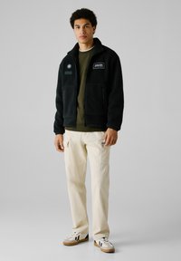 Sort fleecejakke med lynlås og to frontlommer, lagret over en grøn skjorte. Flødefarvede cargo bukser med sidelommer. Casual sneakers.