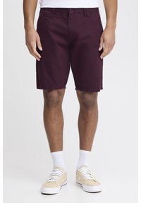 Pantalones cortos marrón de tela ligera, con bolsillos frontales y cierre de botón. Combinados con zapatillas beige y calcetas blancas.