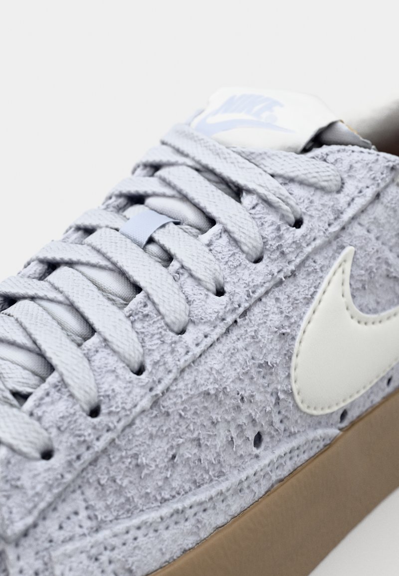 Baskets gris clair avec une tige en daim texturé, un swoosh en cuir blanc, des lacets gris et une semelle en caoutchouc marron. Design minimaliste.