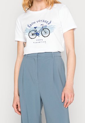 Kobieta ubrana w biały t-shirt z niebieskim rowerem i napisem "Bon Voyage, happy travelling", w połączeniu z niebieskimi spodniami z wysokim stanem.