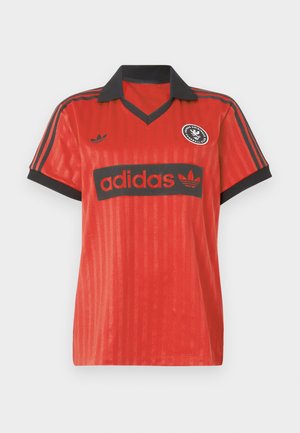 Červený futbalový dres s čiernym golierom, s logom Adidas v čiernej farbe a kruhovou klubovou emblémou na ľavej strane hrudníka.