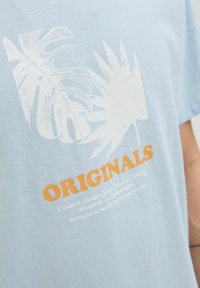 Világoskék pamut póló, fehér trópusi levél nyomattal és narancssárga "ORIGINALS" felirattal, alatta további fehér szöveges részletezéssel.