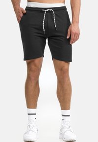 Mann in schwarzen Joggingshorts mit Kordelzug, weißen Kniestrümpfen mit schwarzen Streifen und weißen Sportschuhen vor weißem Hintergrund.