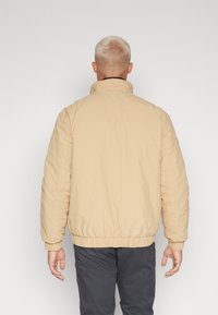 Světle béžová prošívaná bunda s vysokým límcem, elastickými manžetami a páskem. Zobrazuje hladkou, mírně pomačkanou strukturu látky.