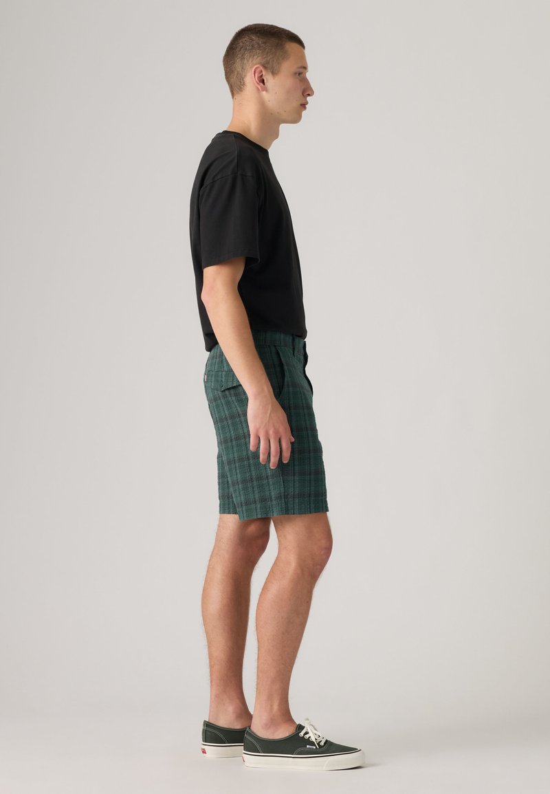 Giovane uomo in piedi di profilo che indossa una t-shirt nera, pantaloncini verdi a quadri e scarpe da ginnastica basse verde scuro con lacci bianchi.