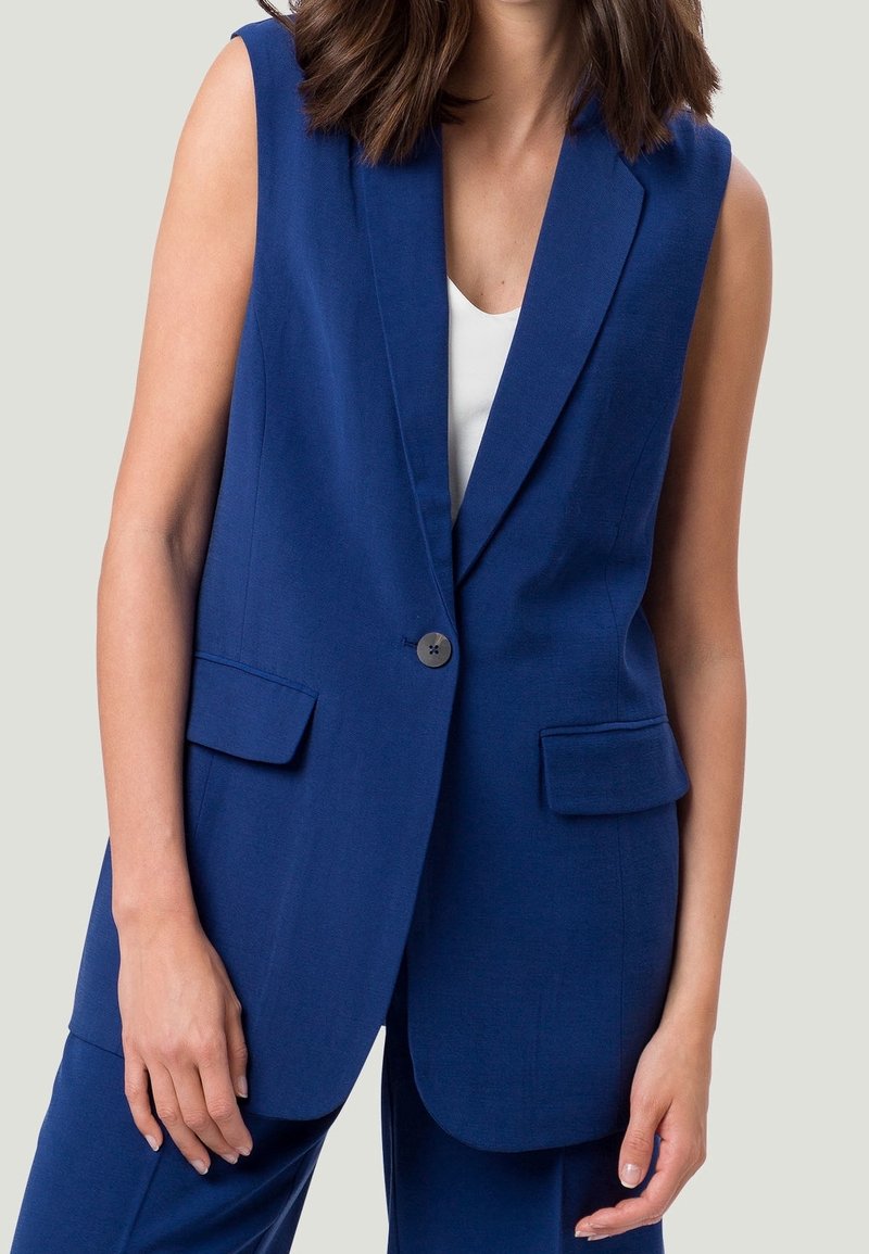 Blazer bleu marine sans manches avec un bouton unique et des poches à rabats frontales, porté sur un haut blanc, vu du cou jusqu'aux hanches.